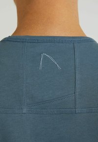 Zadní pohled na osobu oblečenou v modré košili se vyšitým bílým abstraktním logem poblíž výstřihu.