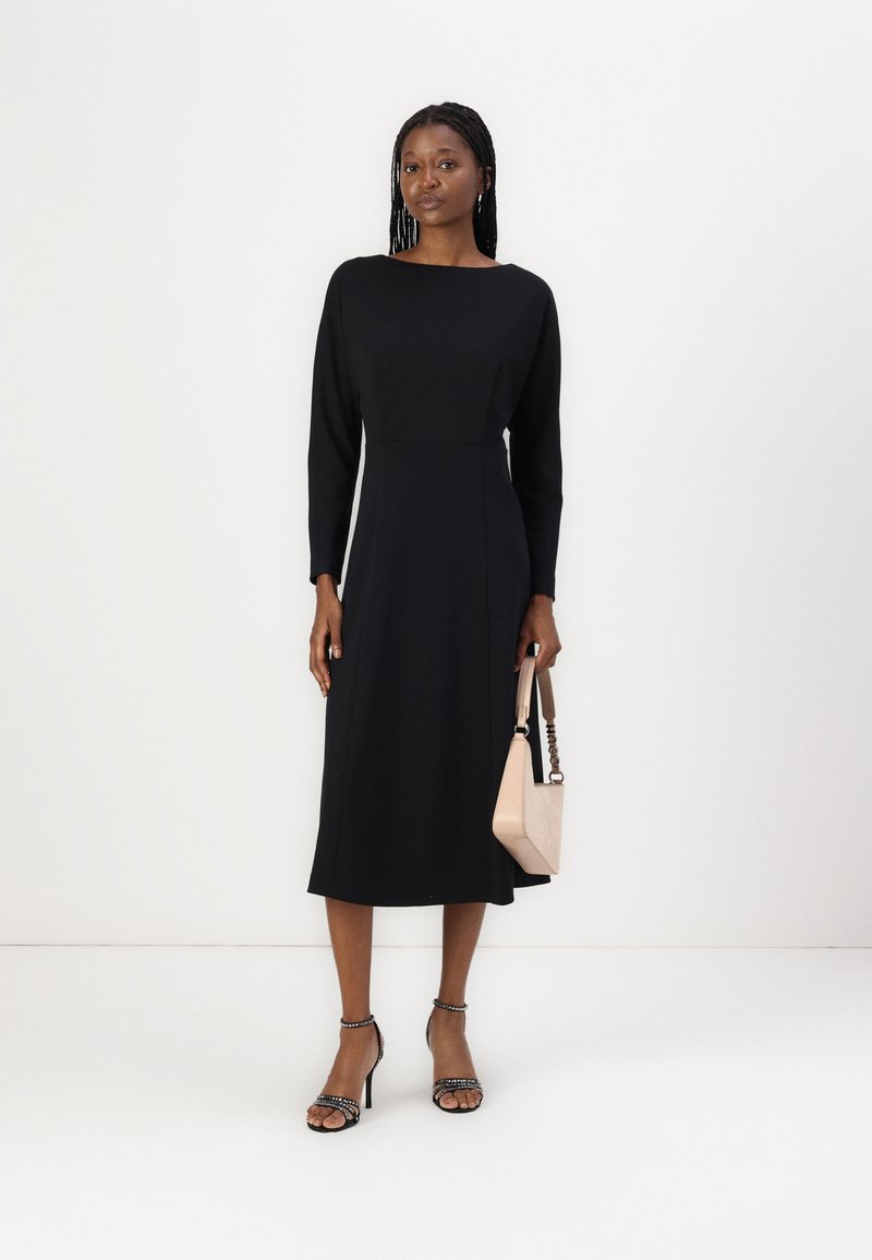 Schwarzes Langarmkleid mit U-Boot-Ausschnitt und taillierter Hüfte, in Midi-Länge. Modell hält eine helle Handtasche und trägt Riemchenabsätze.