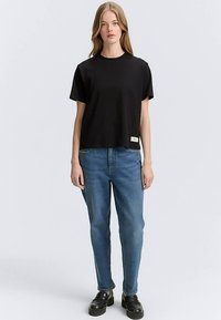 Črna bombažna majica s kratkimi rokavi in okrolim ovratnikom, kombinirana z modrimi kavbojkami iz jeansa in črnimi loafer čevlji, stoji ob nevtralnem ozadju.