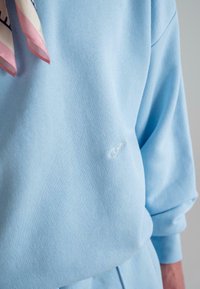 Hellblaues Sweatshirt aus geripptem Stoff, mit einem dezenten, weißen, gestickten "P"-Logo auf der Brust und lockeren Ärmeln.
