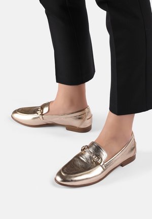 Gouden metallic loafers met een gladde textuur, een ronde neus, decoratieve ringen op de bovenkant en een lage houten hak. Gecombineerd met zwarte broek.