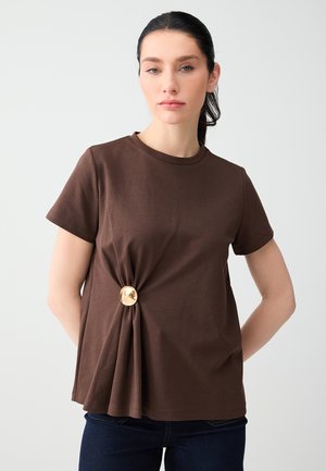 Donna con capelli scuri raccolti indietro che indossa una maglietta marrone a maniche corte con una fibbia decorativa dorata sul lato, in piedi contro uno sfondo chiaro.