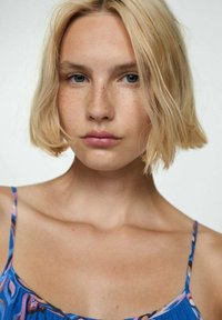 Eine blonde Person mit einem kurzen Bob, die ein blaues Kleid mit bunten Mustern und dünnen Trägern trägt. Sie hat glatte Haut und dezent sichtbare Sommersprossen.