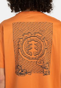 Pomarańczowa, krótko rękawowa koszulka z czarną grafiką przedstawiającą mur z cegieł oraz postać z dymkiem, w którym znajduje się logo na środku.
