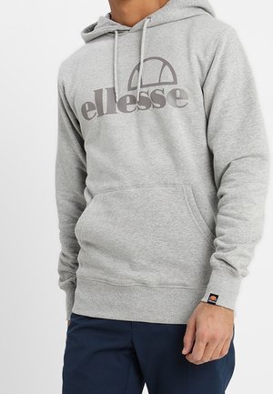 Homme portant un sweat à capuche ellesse gris clair avec une poche avant et un pantalon bleu marine, debout devant un fond uni.