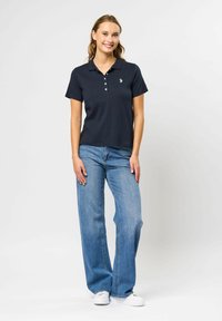 Kvinne smilende stående, iført en svart poloshorte med logo, blå vide jeans og hvite joggesko på ensfarget bakgrunn.