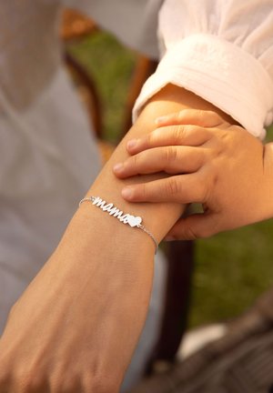 Bracciale d'argento con la parola "mama" e una forma a cuore, posato su un polso mentre una mano di un bambino lo tocca gentilmente.