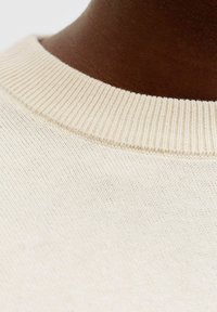 Collo di maglione beige in maglia con texture a coste, vista ravvicinata, che evidenzia il tessuto liscio e la finitura morbida.