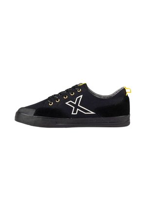 Zapatilla deportiva negra de corte bajo con cordones negros, ojales amarillos, un logo blanco "X" en el lateral y un lazo amarillo en el talón sobre una suela negra.
