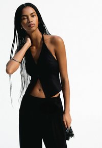 Čierny halter top so skráteným strihom a unikátnym prekrývacím dizajnom, spárovaný s čiernymi nohavicami, ktoré majú čisté línie a hladkú textúru.