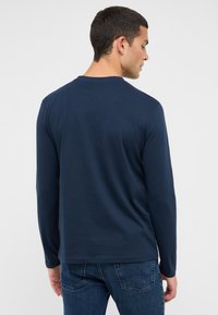 Tumši zils T-krekls ar garām piedurknēm, apaļu kakla izgriezumu, taisnu apakšmalu un mīkstu, gludu tekstūru. Kombinēts ar zilām džinsa biksēm, rādot ērtu piegriezumu.