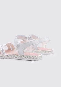 Sandalias gris claro con textura trenzada, tirante en el tobillo y suela blanca con patrón de zigzag. Logo rosa en la plantilla.