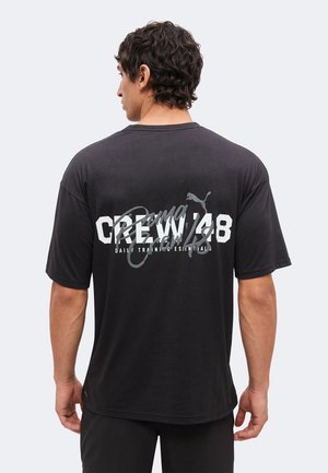 Mand med krøllet mørkt hår iført sort Puma Crew 48 T-shirt med hvid og grå tekst, stående med ryggen mod en lys baggrund.