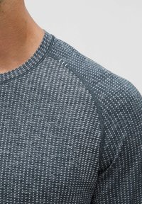 Športové tričko v tmavom teal s textúrovaným vzorom malých štvorcov. Obsahuje rebrovaný okrúhly výstrih a raglánové rukávy.