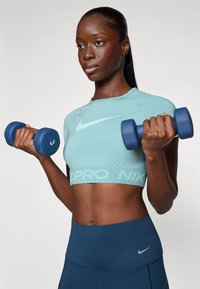 Top atlético corto de color azul claro con banda texturizada y logo, leggings negros, sosteniendo mancuernas azules, contra un fondo liso.