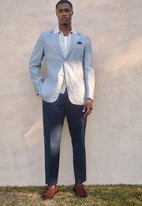 Blazer en lin bleu clair avec boutons dorés, associé à une chemise de couleur claire et un pantalon bleu marine. Mocassins marron, debout sur l'herbe avec un mur texturé en arrière-plan.