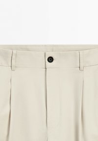 Close-up of beige trousers waistband with black button, belt loops, and front pleats.