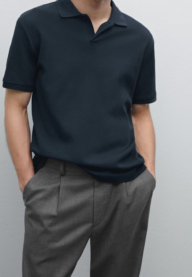 Homme portant un polo à manches courtes bleu marine foncé, légèrement rentré dans un pantalon gris à plis, les mains dans les poches, sur un fond uni.