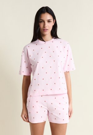 Junge Frau trägt ein hellrosa kurzärmeliges Shirt und passende Shorts mit kleinen roten Rosenmustern vor einfachem beigem Hintergrund.