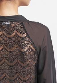 Zwarte blouse van doorschijnende stof met ingewikkelde kantdetails op de rug. Het kant toont een golvend patroon met bloemmotieven.