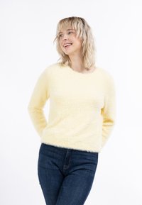 Puhaszőrű sárga pulóver kerek nyakkivágással és hosszú ujjakkal, sötétkék farmernadrággal párosítva. A szövet lágy textúrája jól látható.