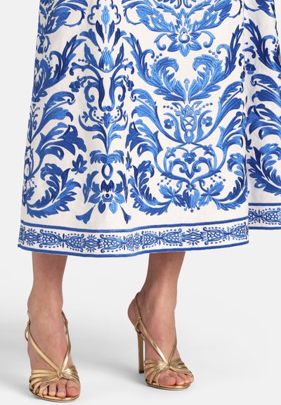 Biela sukňa s detailnou modrou kvetinovou výšivkou nad zlatými sandálmi na vysokom opätku s remienkami na bosých nohách na bielej podklade.