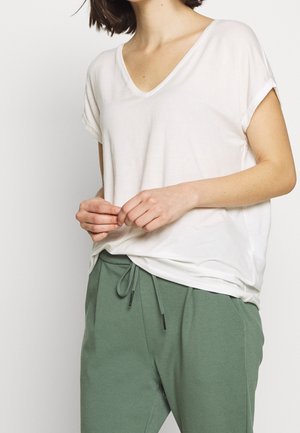 Biała koszulka z dekoltem w serek i podwiniętymi krótkimi rękawami, zestawiona z zielonymi spodniami na sznurek. Materiał wydaje się miękki i o luźnym kroju.