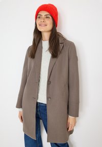 Cappotto di lana marrone con colletto a risvolto, due bottoni e tasche laterali, indossato sopra un maglione grigio chiaro e jeans blu. Berretto di maglia rosso sulla testa.