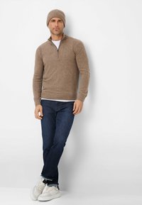 Hellbrauner Strickpullover mit Reißverschlusskragen, getragen über einem weißen Hemd, kombiniert mit dunkelblauen Jeans und weißen Sneakers, abgerundet mit einer passenden Mütze.