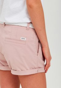 Rosa bomullsshorts med rullade fållar, med en liten bakficka och en etikett. Bärs med ett vitt flätat bälte.