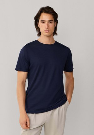 Hombre joven con cabello castaño de longitud media, vestido con una camiseta de manga corta azul marino y pantalones beige claros, de pie contra un fondo liso.