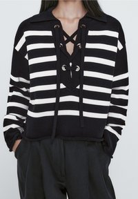 Pull en rayures noir et blanc avec col, devant à lacets doté d'œillets en métal, et coupe décontractée. Tissu tricoté texturé.