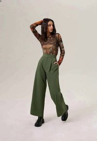 Long-sleeved, patterned top in brown with artwork design; high-waisted, wide-leg green trousers; black ankle boots; model posing against a light background.