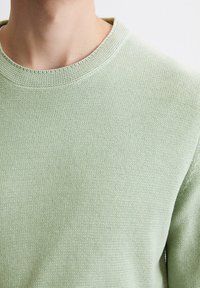 Hellgrüner gestrickter Pullover mit einem gerippten Rundhalsausschnitt und strukturiertem Stoff. Nahaufnahme zeigt eine glatte Oberfläche mit subtilen Stichmustern.