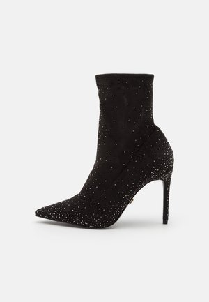 Schwarzer Wildleder-Ankle-Boot mit spitzem Zeh, verziert mit verstreuten metallischen Nieten und einem schlanken hohen Absatz. Weiche Textur und figurbetonten Design.