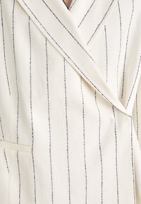 Blazer crema con cuello chal, con rayas verticales negras, diseño de doble botonadura y detalle de bolsillo frontal. Tejido texturizado.