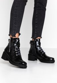 Bottines noires en vernis avec une texture crocodile, lacets à l'avant, quincaillerie argentée et un talon chunky, associés à un jean noir effiloché.