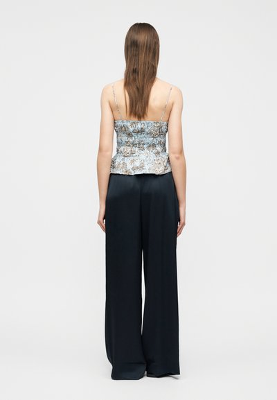 Γυναίκα με μακριά καστανά μαλλιά που φοράει μπλε φλοράλ αμάνικο τοπ και άνετο μαύρο παντελόνι φαρδιάς γραμμής, standing με την πλάτη προς το φακό σε απλό φόντο.