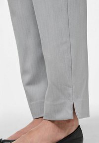 Pelēki piegriezti bikses ar gludu tekstūru, ar sānu šķēlumiem apakšmalē un kombinētas ar melnu apavu.