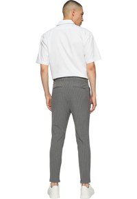 Homme portant une chemise blanche à manches courtes, pantalon slim gris à fines rayures et baskets blanches, debout de dos sur un fond blanc uni.