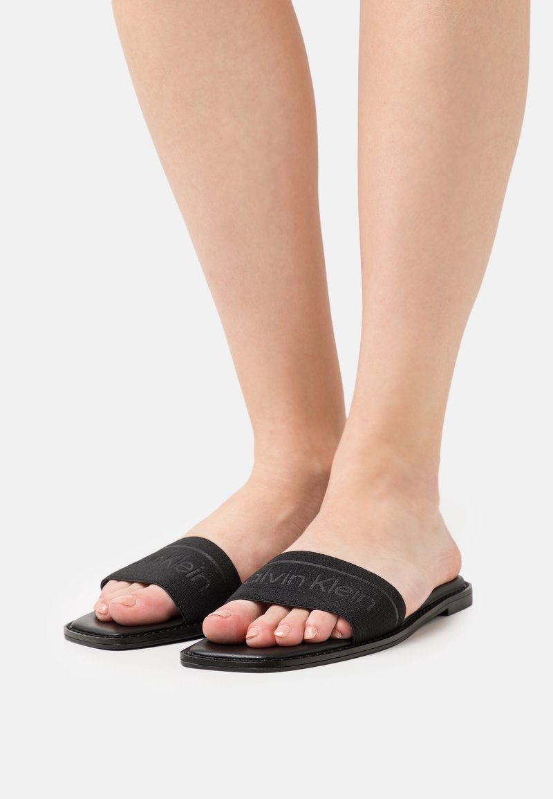 Personne portant des sandales slides Calvin Klein noires à bout ouvert avec une semelle plate sur un fond clair uni.