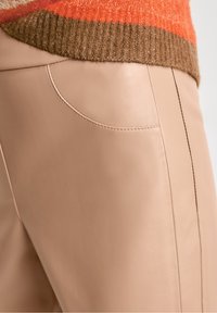 Beige läderbyxor med en slät yta, som har en sidoficka och rena sömmar. Toppen visar en texturerad orange och brun stickad tröja.