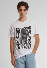 Balta kokvilnas T-krekls ar melnu grafisko druku, kurā attēlots basketbola spēlētājs, un distorted text "MANI SPĒLE NEDĒĻAI". Standarta piegriezums.
