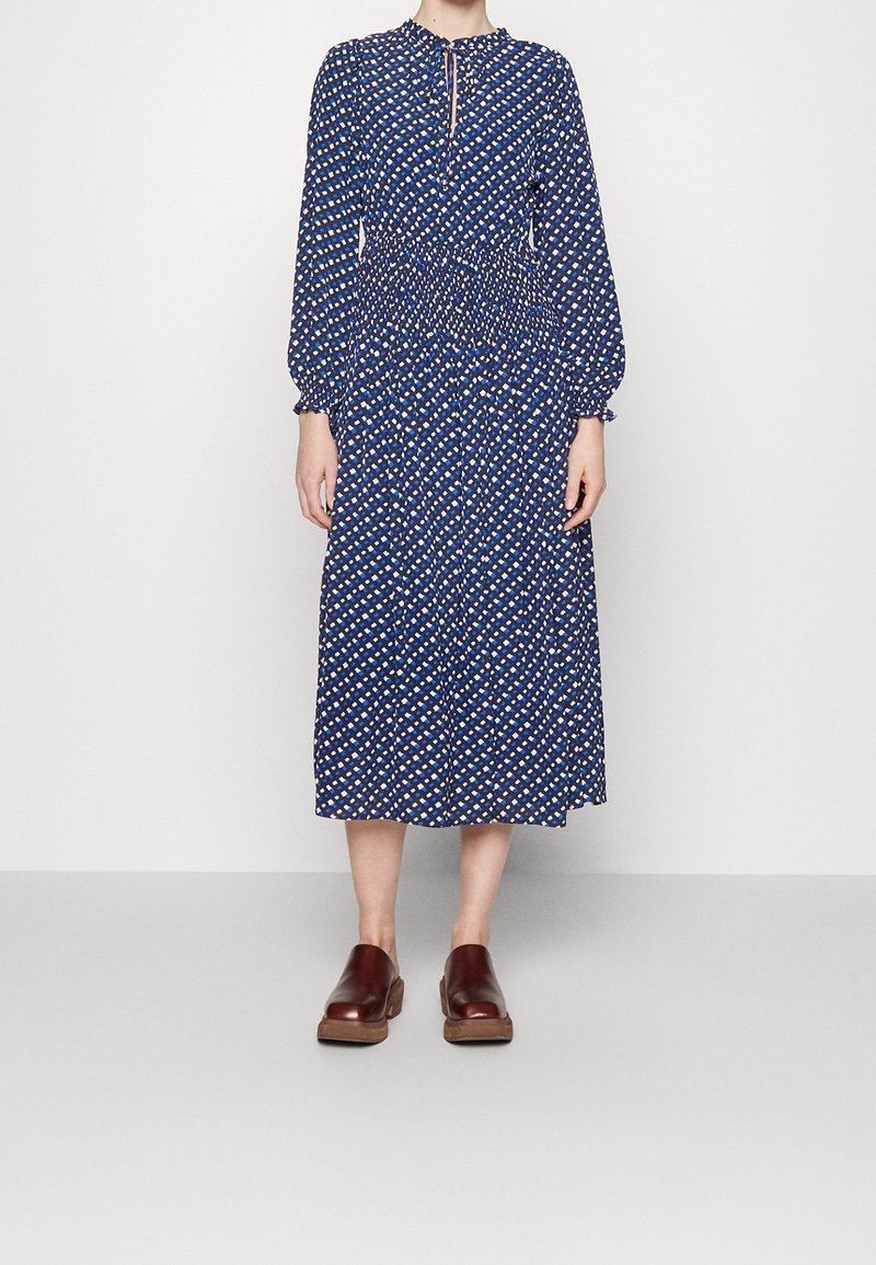 Robe midi bleu marine avec un motif en losanges, taille smockée, longues manches bouffantes et détail de décolleté en œillet. Assortie avec des sabots marron.