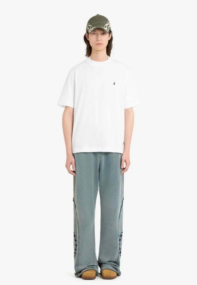 Jeune personne portant un T-shirt blanc oversized, un pantalon ample gris délavé avec des motifs d'étoiles, des chaussures marron, et une casquette vert olive ornée de motifs de crânes.