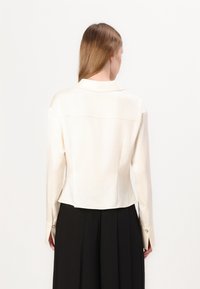 Blusa in seta avorio con maniche lunghe, vestibilità slim e colletto a punte. Presenta bottoni dorati sui polsini e una texture liscia e lucente.