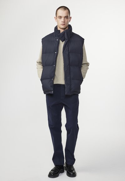 Marineblaues, Steppweste mit hohem Kragen und Druckknöpfen. Getragen über einem hellbeigen Pullover mit strukturiertem Strick, kombiniert mit dunklen Hosen.