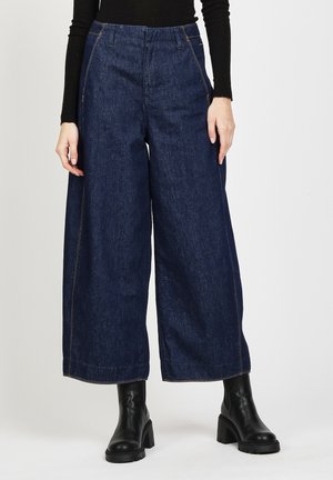 Weite Denim-Hosen in Dunkelblau mit gelben Nähten. Sie haben eine hohe Taille und einen geraden Schnitt. Kombiniert mit schwarzen Knöchelstiefeln.