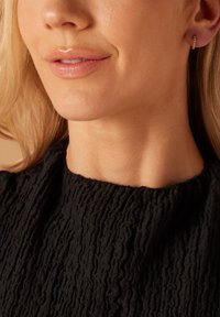 Boucle d'oreille en or avec des accents à pois, portée sur le lobe de l'oreille. Haut en tissu noir texturé avec un design froncé, gros plan du visage et du cou.