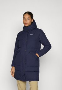 Marineblauwe gewatteerde jas met een hoge kraag, met drukknopen aan de voorkant, twee zijzakken en textuurstof; heeft een logo op de linkerkant van de borst.